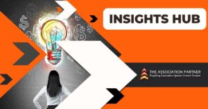 The featured image for 'Insights Hub' displays a creative and vibrant design. At the center is a large, brightly colored light bulb with a brain-like illustration within it, symbolizing innovative thinking and ideas. In front of the light bulb stands a professional woman in a white shirt, viewed from the back, looking up at the light bulb, indicative of contemplation and vision. The background is a mix of gray with doodles of currency symbols and arrows, implying a strategic approach to financial success. The upper right corner has a bold, orange banner with the words 'INSIGHTS HUB' in white, next to the logo of 'THE ASSOCIATION PARTNER' which includes a tagline 'Propelling Associations Upward. Onward. Forward.' The overall design conveys a theme of dynamic business strategy and intellectual growth.